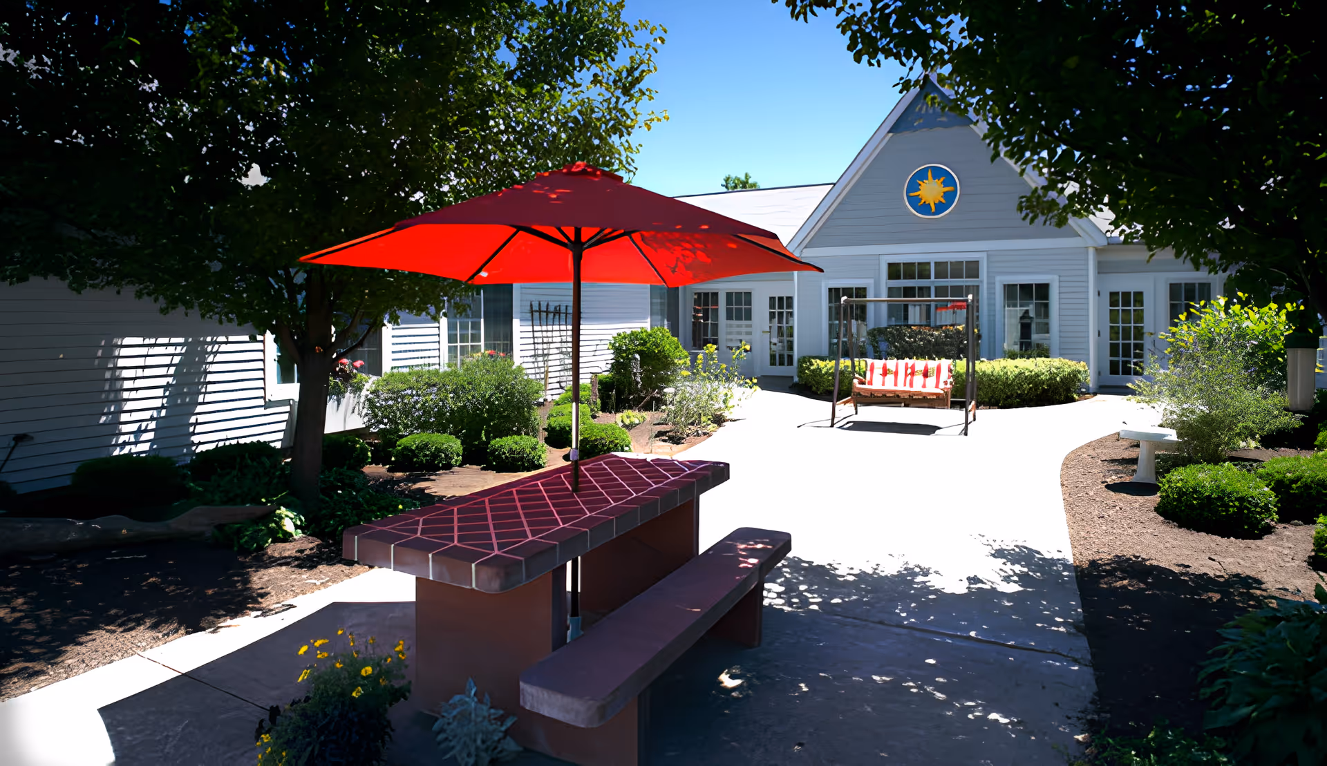 Outdoor courtyard area with a red umbrella over a tiled picnic table and bench, surrounded by trees and bushes. In the background, there is a white building with multiple windows and doors, a swing with red and white cushions, and a clear blue sky.