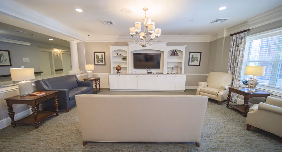 A well-lit living room area in a senior living facility featuring a beige sofa facing a white entertainment center with a flat-screen TV. The room includes a blue couch, two beige armchairs, wooden side tables with lamps, framed artwork on the walls, a chandelier, and large windows with patterned curtains allowing natural light to enter.