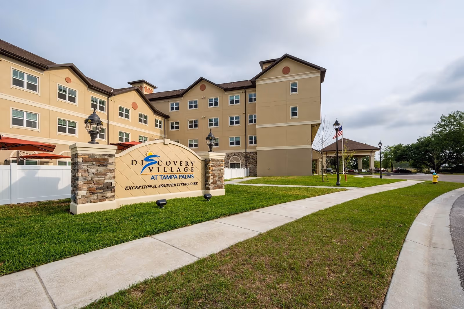 Exterior view of Discovery Village At Tampa Palms, a multi-story assisted living facility with beige walls and multiple windows. In front of the building is a large sign with the facility's name and the phrase 'Exceptional Assisted Living Care.' The area features a well-maintained lawn, a sidewalk, street lamps, and an American flag.