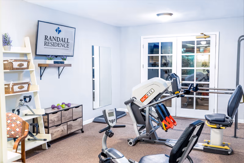 Small fitness room with exercise machines, an exercise bike, free weights, shelving and a wall-mounted sign reading 'Randall Residence'.