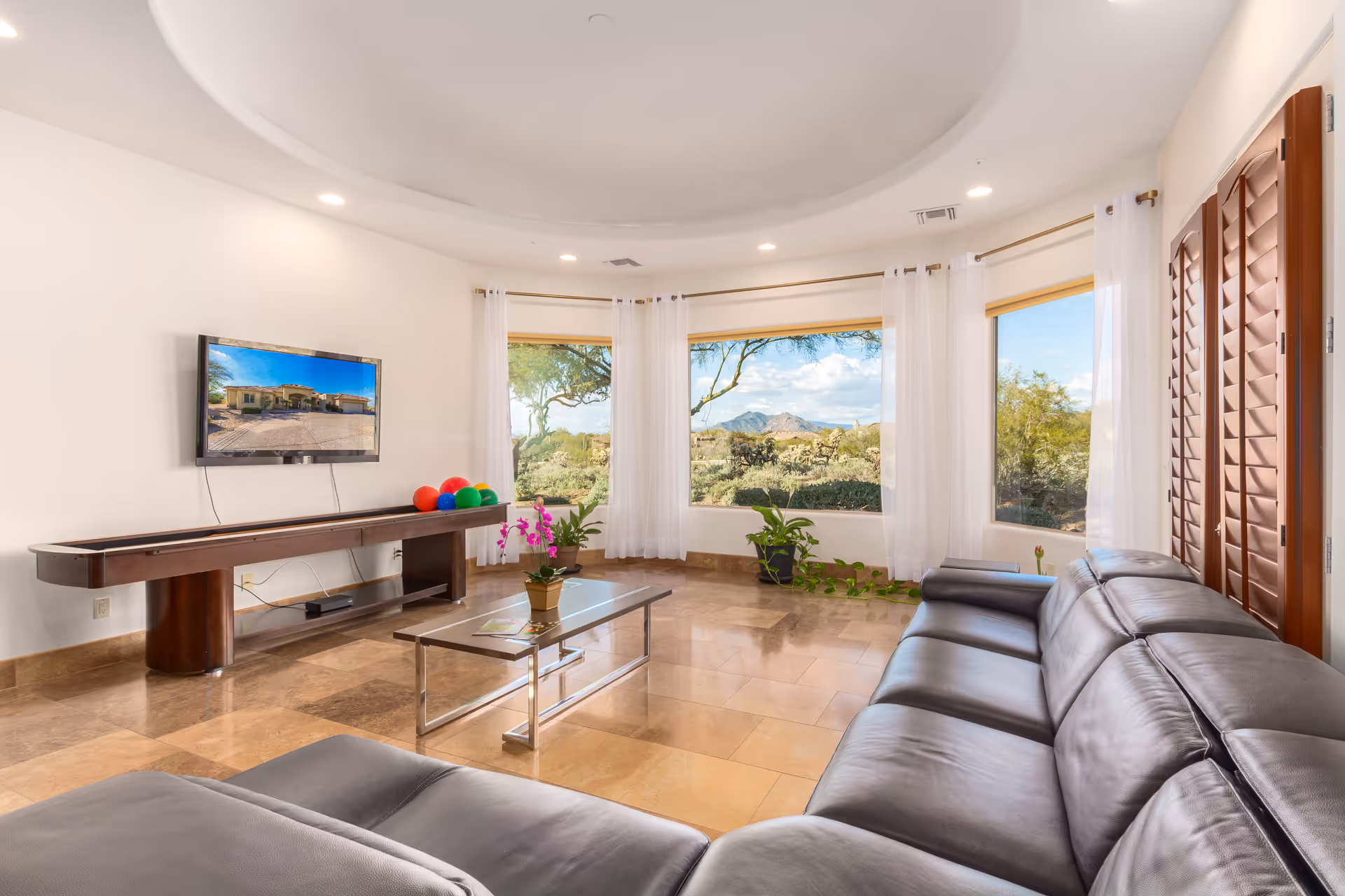Bright living room with a large leather sectional facing a wall-mounted TV, glass coffee table, shuffleboard table and large windows showing desert views.
