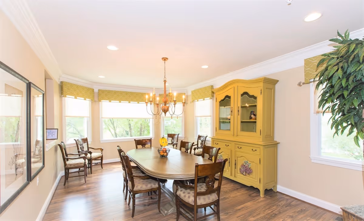 Bright dining room with a long wooden table, chandelier, multiple chairs, and a yellow china cabinet by windows.