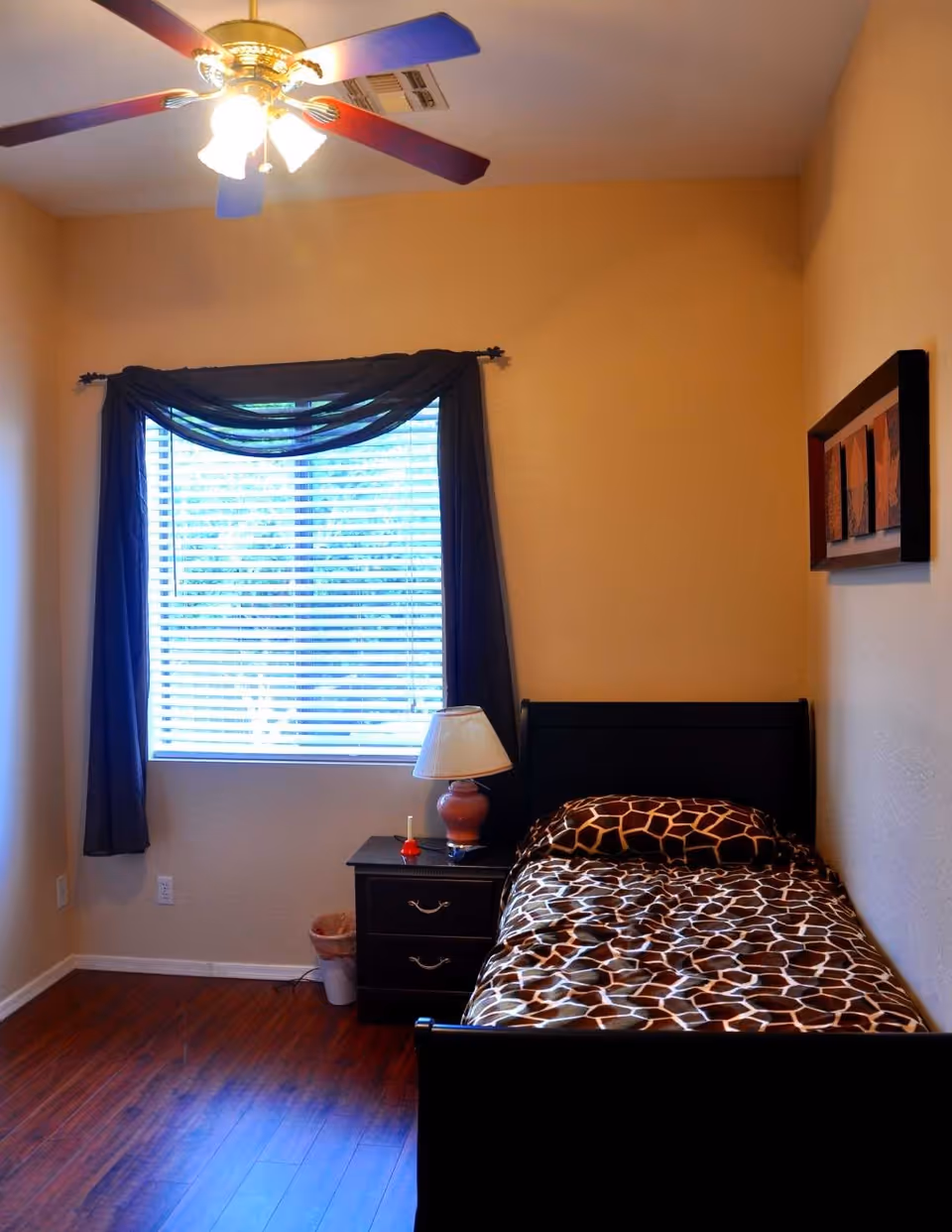 A small bedroom with a single bed covered in giraffe print bedding, a black nightstand with a lamp and small items, a window with blinds and dark curtains, a ceiling fan with lights, and a framed artwork on the wall.