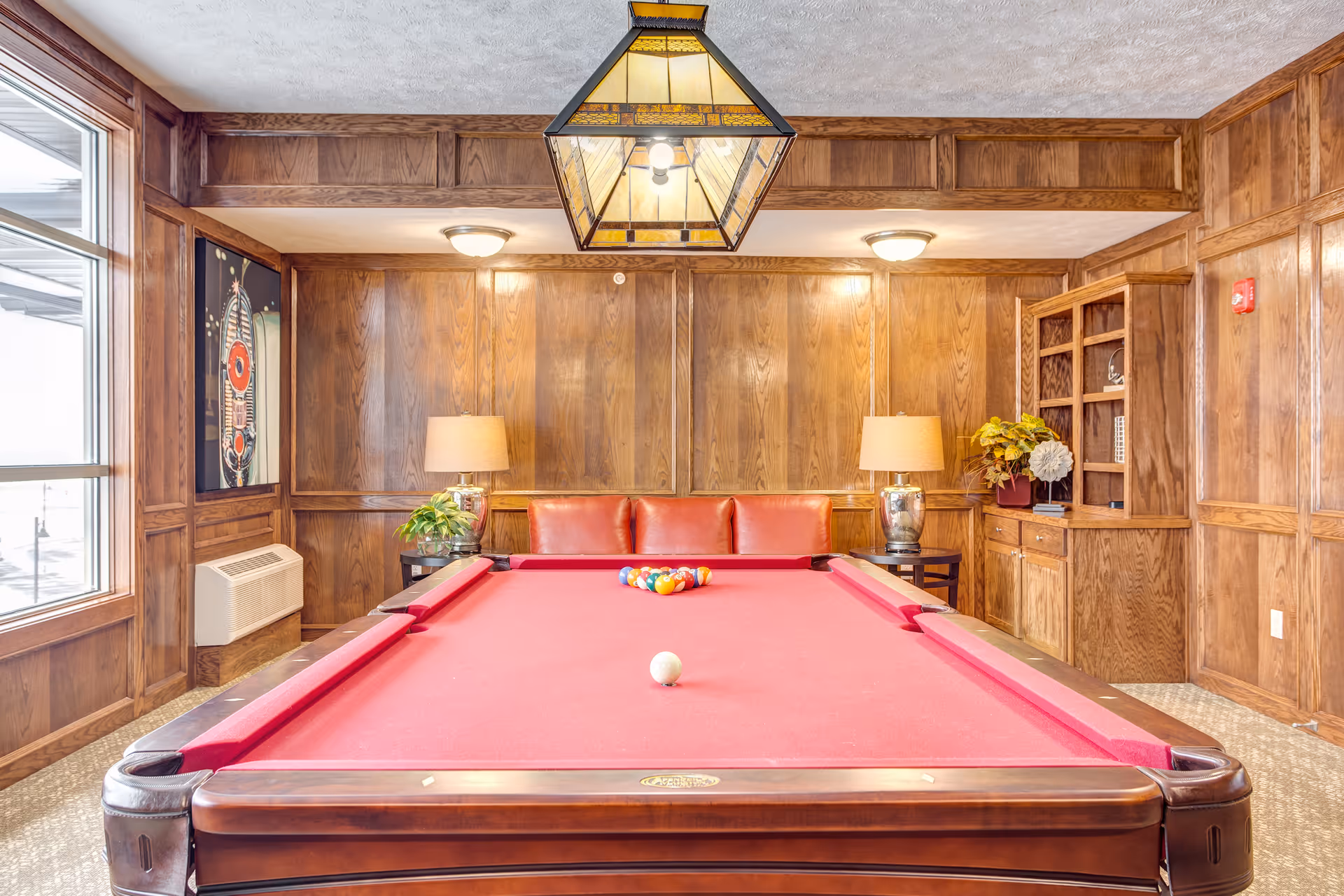 A cozy game room with a red felt pool table in the center, a set of billiard balls arranged at one end, and a white cue ball near the other end. The room features wood-paneled walls, a large window on the left, two table lamps on either side of a red leather couch against the back wall, and a built-in wooden cabinet with shelves on the right. A stained glass pendant light hangs above the pool table.