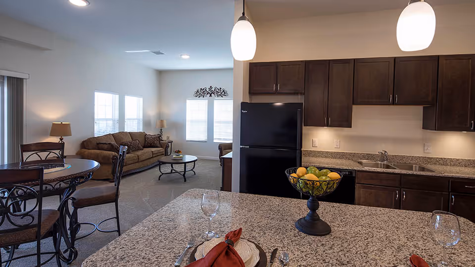 View of an open-concept living area in a senior apartment showing a kitchen with granite countertops, dark wood cabinets, a black refrigerator, and a double sink. The kitchen island has a decorative fruit bowl and place settings with wine glasses. Adjacent to the kitchen is a dining area with a round table and chairs, and a living room with a brown sofa, coffee table, and windows letting in natural light.
