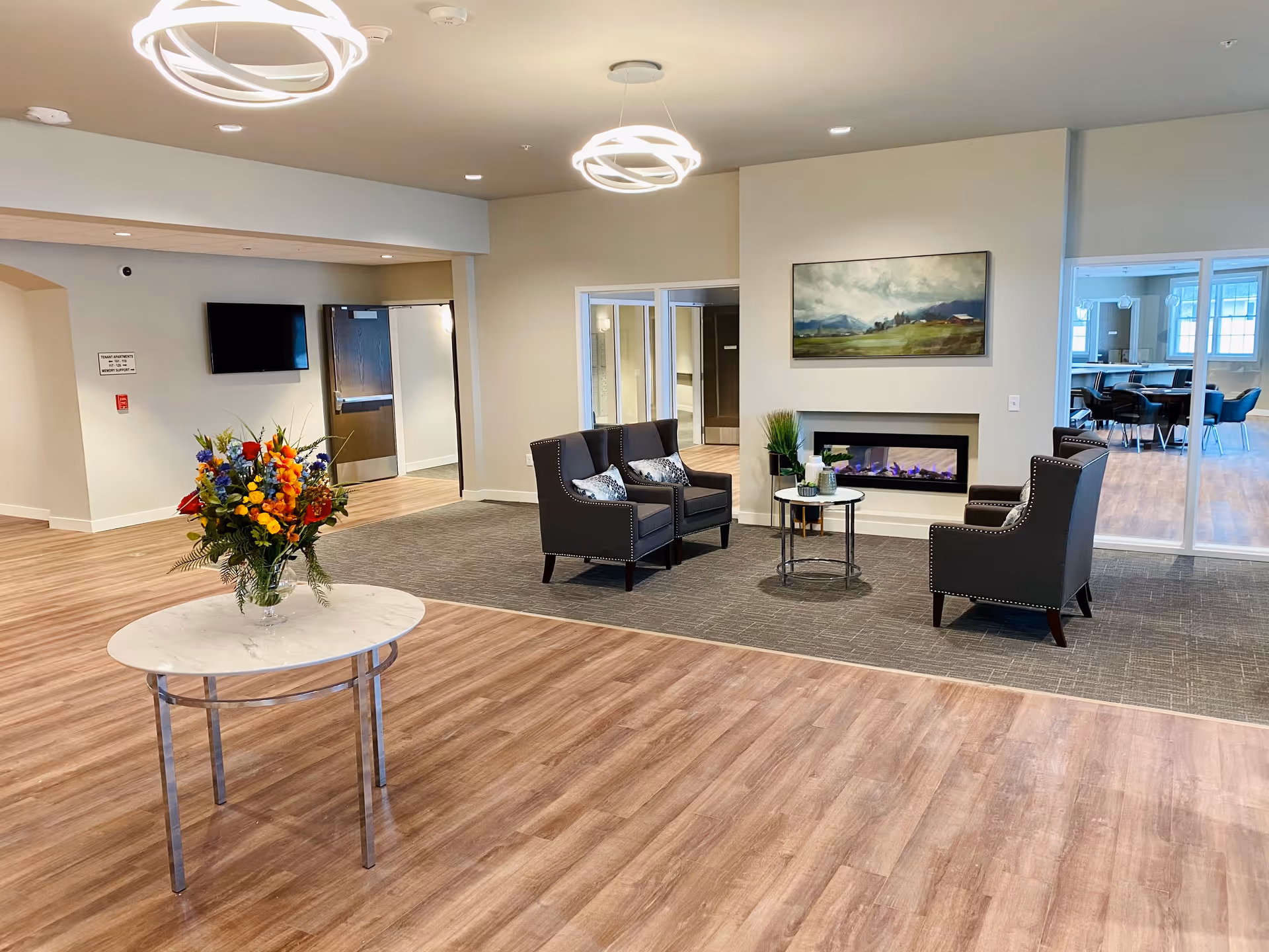 A spacious and well-lit assisted living facility common area with wood flooring and carpeted seating area. The seating area includes three dark upholstered armchairs arranged around a small round table with decorative items. Above the seating area is a landscape painting and a modern electric fireplace. In the foreground, there is a round marble table with a colorful flower arrangement. The background shows a hallway with a mounted TV and a door, as well as a dining area with tables and chairs visible through glass doors.