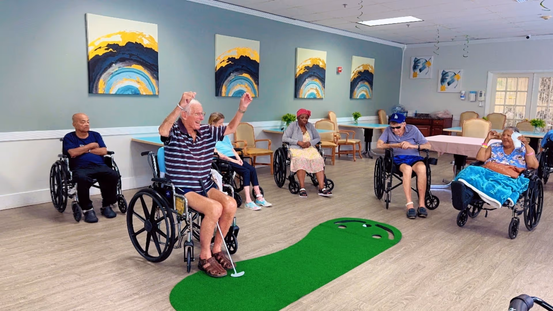 A group of elderly individuals, some in wheelchairs, participating in an indoor putting golf activity in a spacious room with light-colored walls and wooden flooring. The room is decorated with abstract paintings and has tables and chairs arranged around the perimeter.
