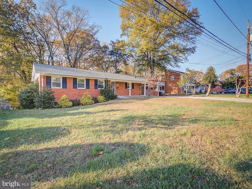 A single-story red brick residential building with a gray roof, surrounded by a grassy lawn and trees with autumn foliage under a clear blue sky. There are power lines overhead and neighboring houses visible in the background.
