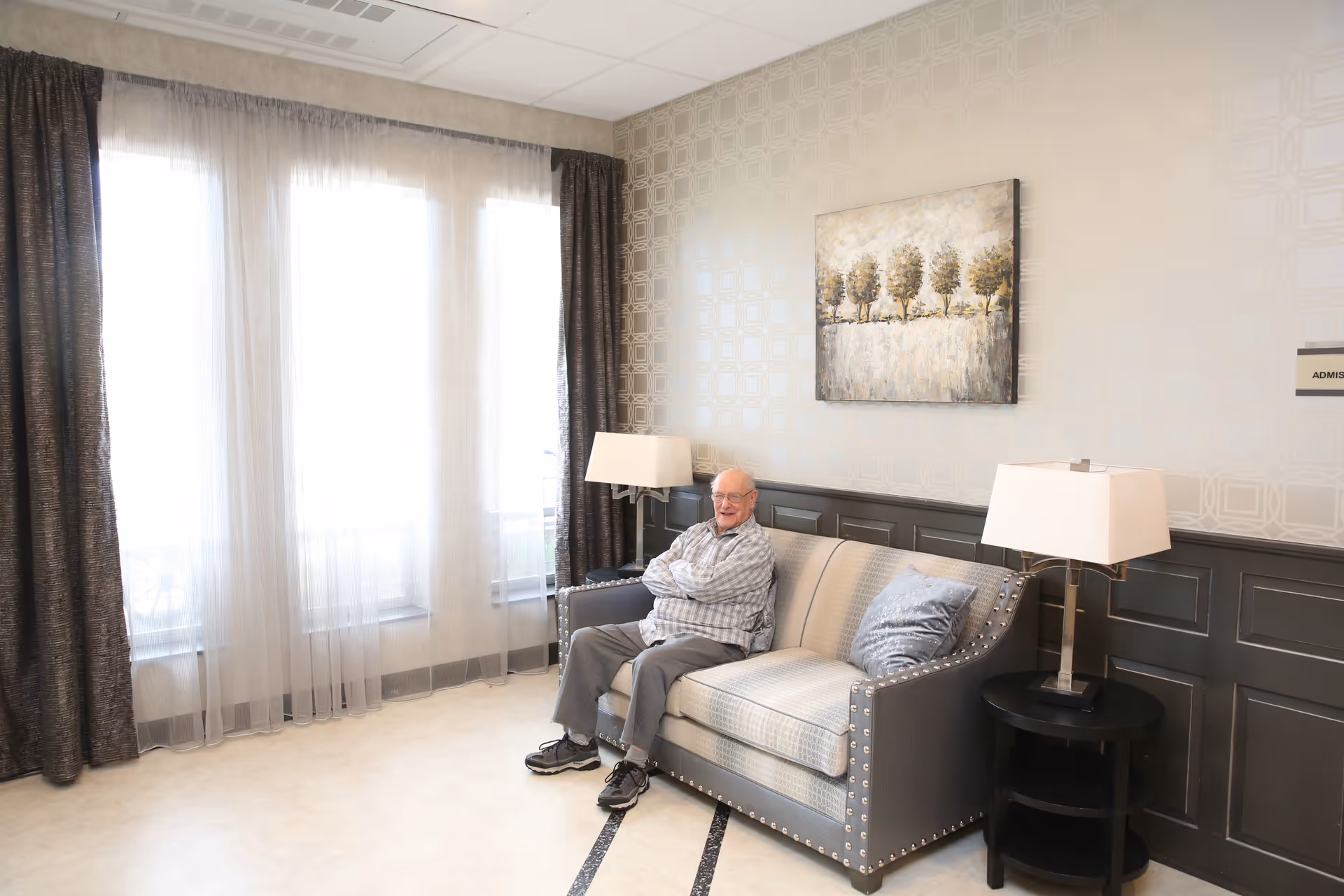 An elderly man sitting on a gray and beige couch in a well-lit room with large windows covered by sheer white curtains and dark drapes. The room has patterned wallpaper, two table lamps on either side of the couch, a small round black side table, and a painting of trees on the wall behind the man.