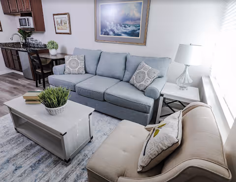 Bright furnished living room with a light blue sofa, beige armchair, coffee table, lamp, and a small kitchenette visible in the background.