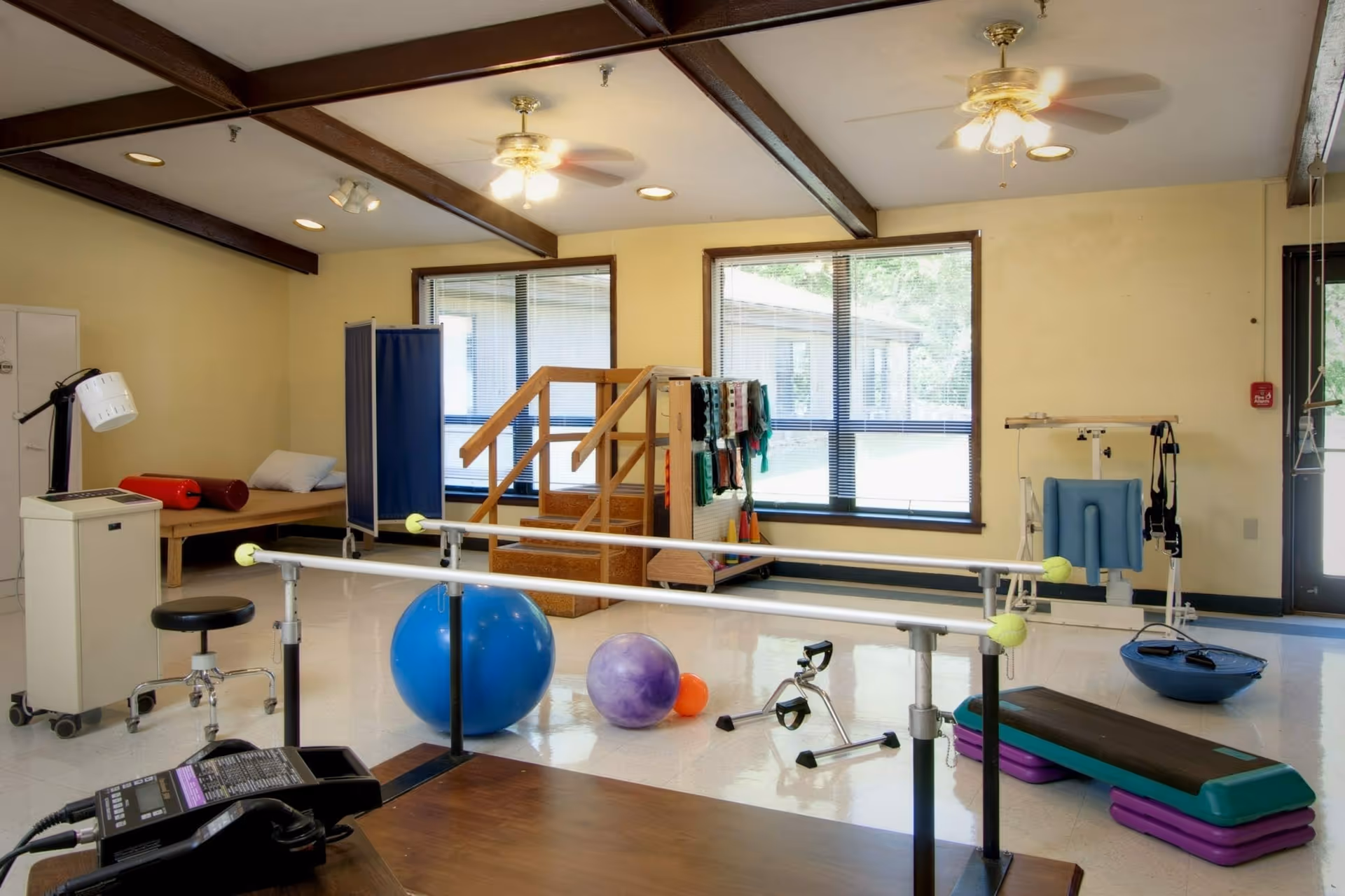 Physical therapy room with parallel bars, exercise balls, step platform, exercise bike, and other rehabilitation equipment. The room has large windows with blinds, ceiling fans, and wooden beams on the ceiling.