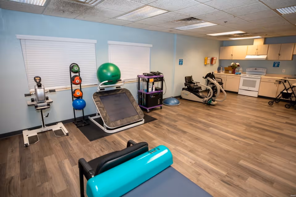A room with exercise equipment including a rowing machine, medicine balls on a rack, a large green exercise ball, a trampoline, a recumbent exercise bike, and a balance trainer. The room has light blue walls, wood flooring, two windows with white blinds, and a kitchenette area with cabinets, a stove, and a walker.