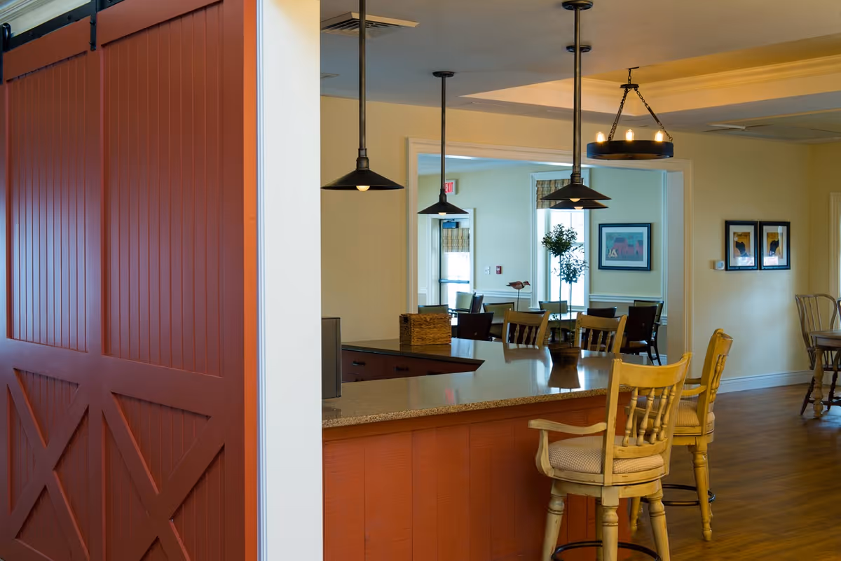 Interior view of a senior living facility dining area with a large countertop island and wooden bar stools. The space features hanging pendant lights, framed artwork on the walls, and a dining table with chairs in the background. A red sliding barn door is partially visible on the left side.