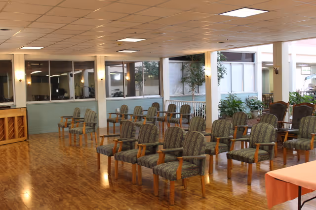 A spacious room with multiple rows of cushioned chairs with wooden arms arranged facing a piano in the corner. The room has large windows, wood flooring, and several plants near the windows, creating a bright and welcoming atmosphere.