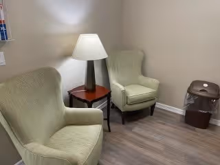A small seating area with two light green upholstered armchairs positioned around a wooden side table with a lamp on it. The floor is wood laminate, and there is a trash can with a liner in the corner against a beige wall.