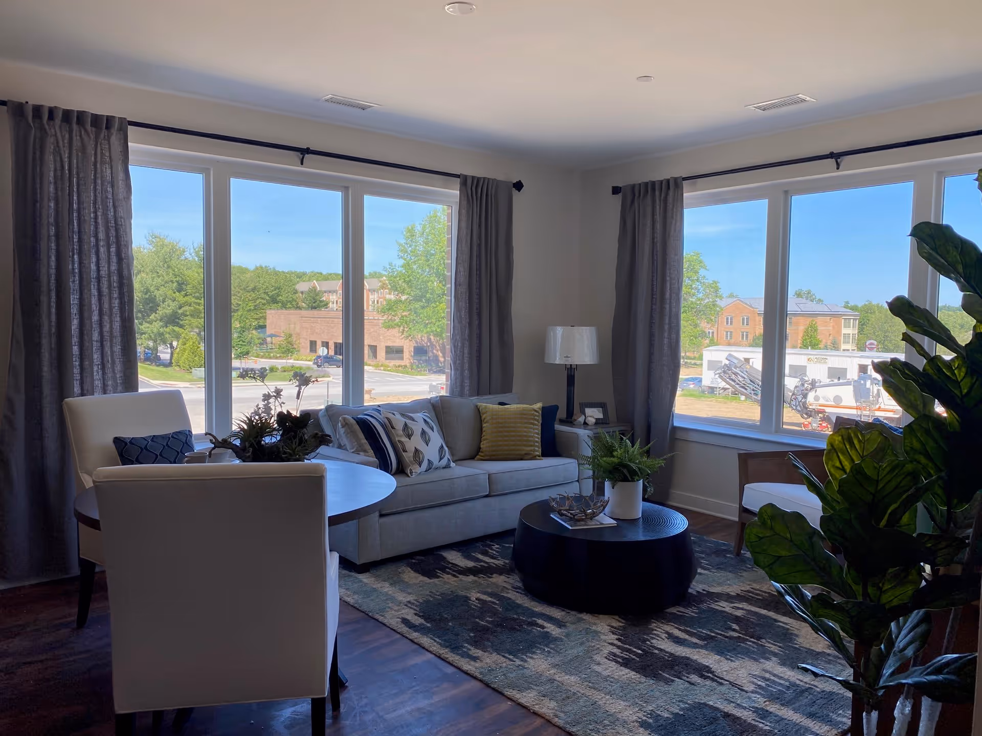 Sunlit living room with a sofa, chairs, round coffee table, rug, and large windows overlooking the street.