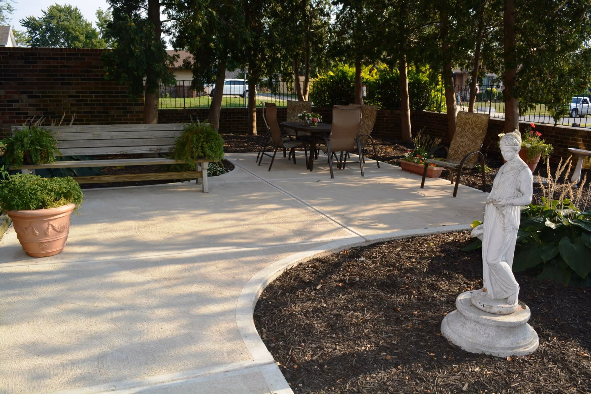 Outdoor patio area with a concrete walkway, a wooden bench with potted plants on either side, a round table with four chairs, and a white statue of a woman standing on a pedestal surrounded by mulch and plants. Trees and a brick wall with a metal fence are in the background.