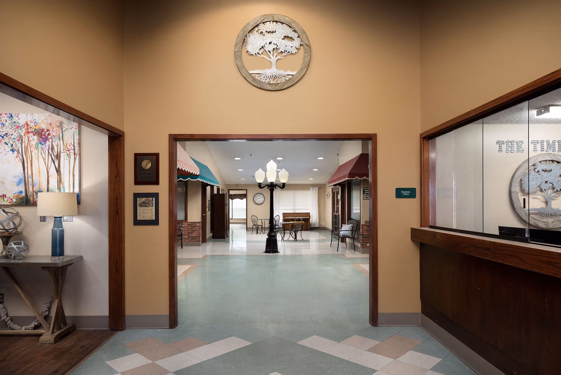 Interior view of a senior living facility hallway with a decorative tree wall art above the doorway. The hallway leads to a common area with tables, chairs, and a lamp post-style light fixture. On the left side, there is a table with a lamp and decorative items, and on the right side, there is a reception window with the facility name 'The Timbers Skilled Nursing & Therapy' visible on the wall behind it.