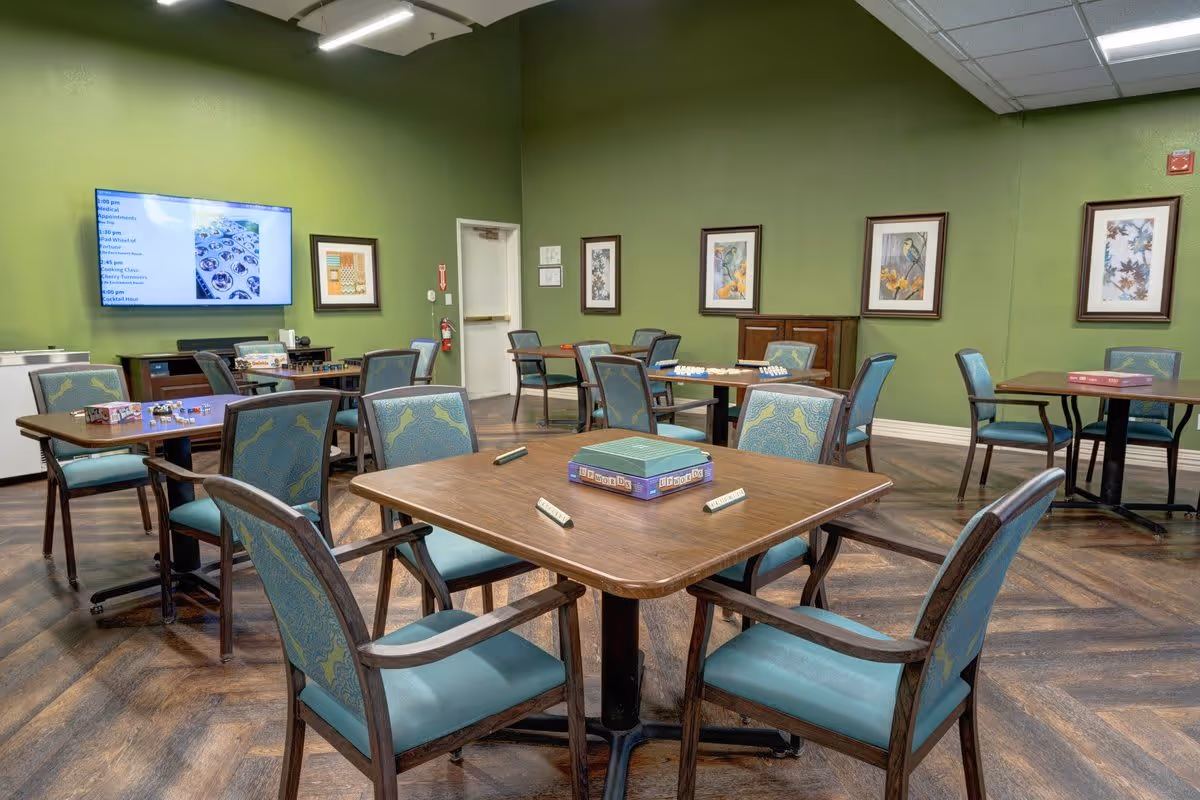 A community room with several square tables and blue cushioned chairs arranged around them. Board games and game pieces are placed on the tables. The walls are painted green and decorated with framed artwork. A large flat-screen TV is mounted on one wall displaying a schedule. The floor has a wood pattern.