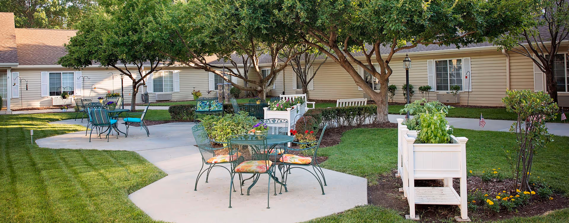 Outdoor courtyard area at Bickford of Lincoln featuring green metal tables and chairs with colorful cushions, white planters with greenery, trees providing shade, and a beige building with white window shutters in the background.