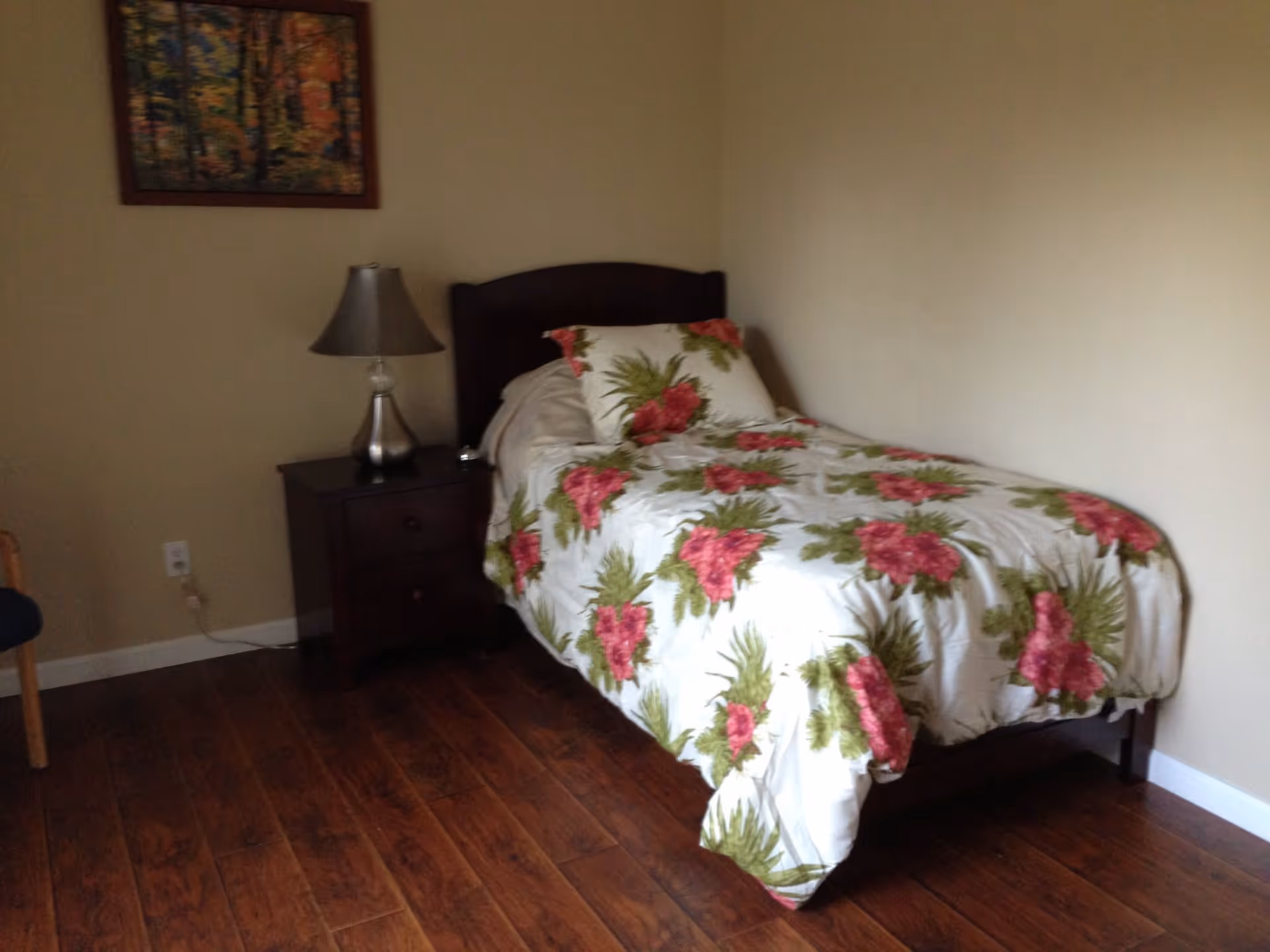 A small bedroom with a single bed covered in a white bedspread with pink and green floral patterns. Next to the bed is a dark wooden nightstand with a silver lamp on it. The room has wooden flooring and cream-colored walls with a framed picture hanging above the nightstand.