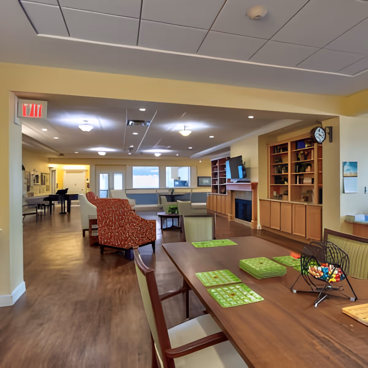 A spacious senior living common area with a wooden table in the foreground set with green placemats and a decorative centerpiece. The room features comfortable seating including patterned armchairs and sofas, a fireplace with a mounted TV above it, built-in wooden bookshelves, and a piano in the background. The walls are painted yellow and the floor has a wood finish.