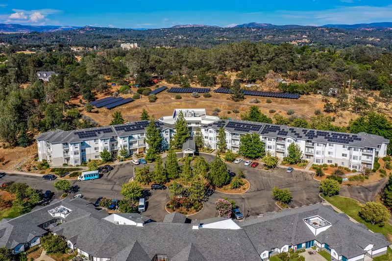 Aerial view of the Country Crest Senior Living complex showing multiple white residential buildings, solar panels on roofs, a circular driveway and surrounding trees and hills.