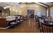 Spacious dining area in a senior living community featuring wooden floors, multiple tables and chairs, a large island with bar stools, pendant lighting, and ceiling fans. The room has a warm, inviting atmosphere with beige walls and large windows allowing natural light.