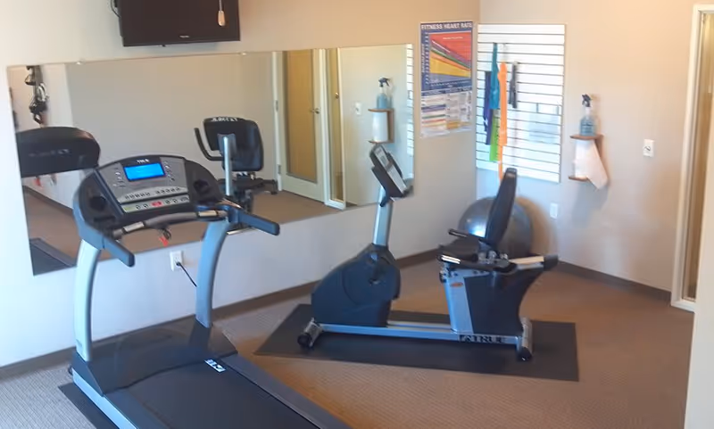 A small fitness room with a treadmill and a recumbent exercise bike on mats. There is a large mirror on the wall behind the equipment, a fitness heart rate chart, resistance bands hanging on a slat wall, a stability ball, and a spray bottle with paper towels mounted on the wall.