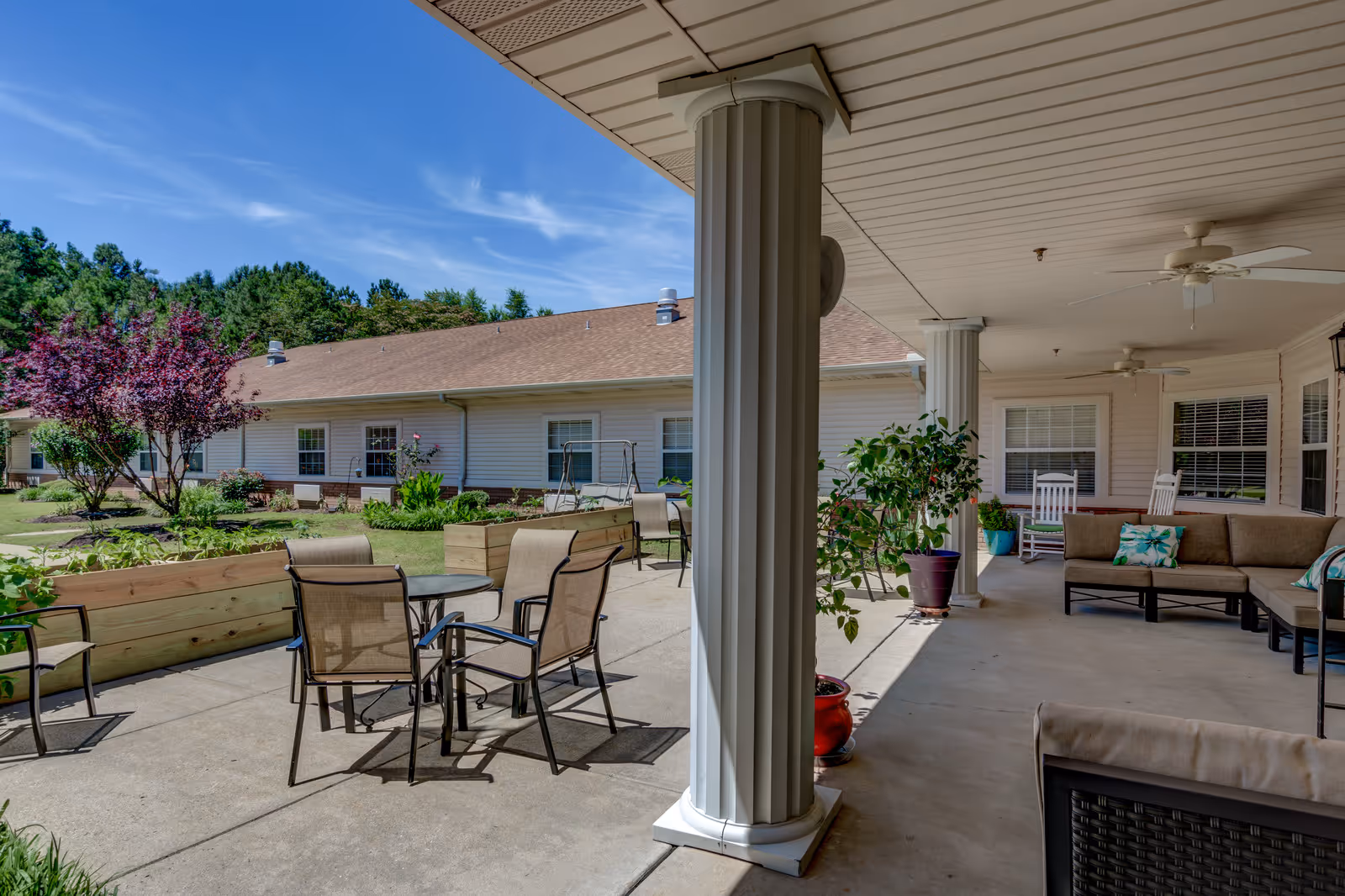 Covered patio at a senior living community with columns, outdoor seating, potted plants, and raised garden beds facing a one-story building.