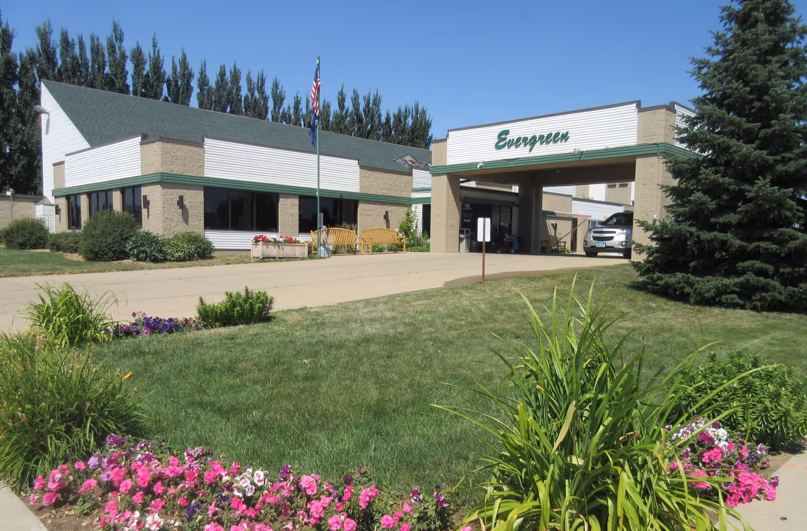 Exterior view of the Evergreen senior living facility showing a one-story building with beige brick and white siding, green roof trim, and a covered entrance. There is a driveway leading to the entrance, a flagpole with an American flag, benches, and well-maintained landscaping with green grass, bushes, and colorful flowers.