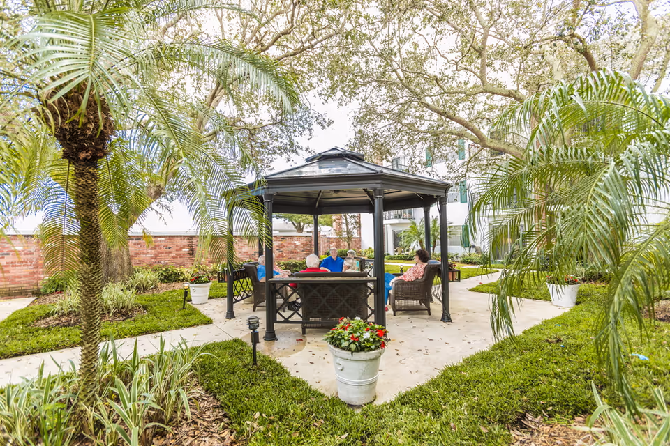 People sitting under a gazebo in a landscaped courtyard with palm trees and potted plants next to a building.