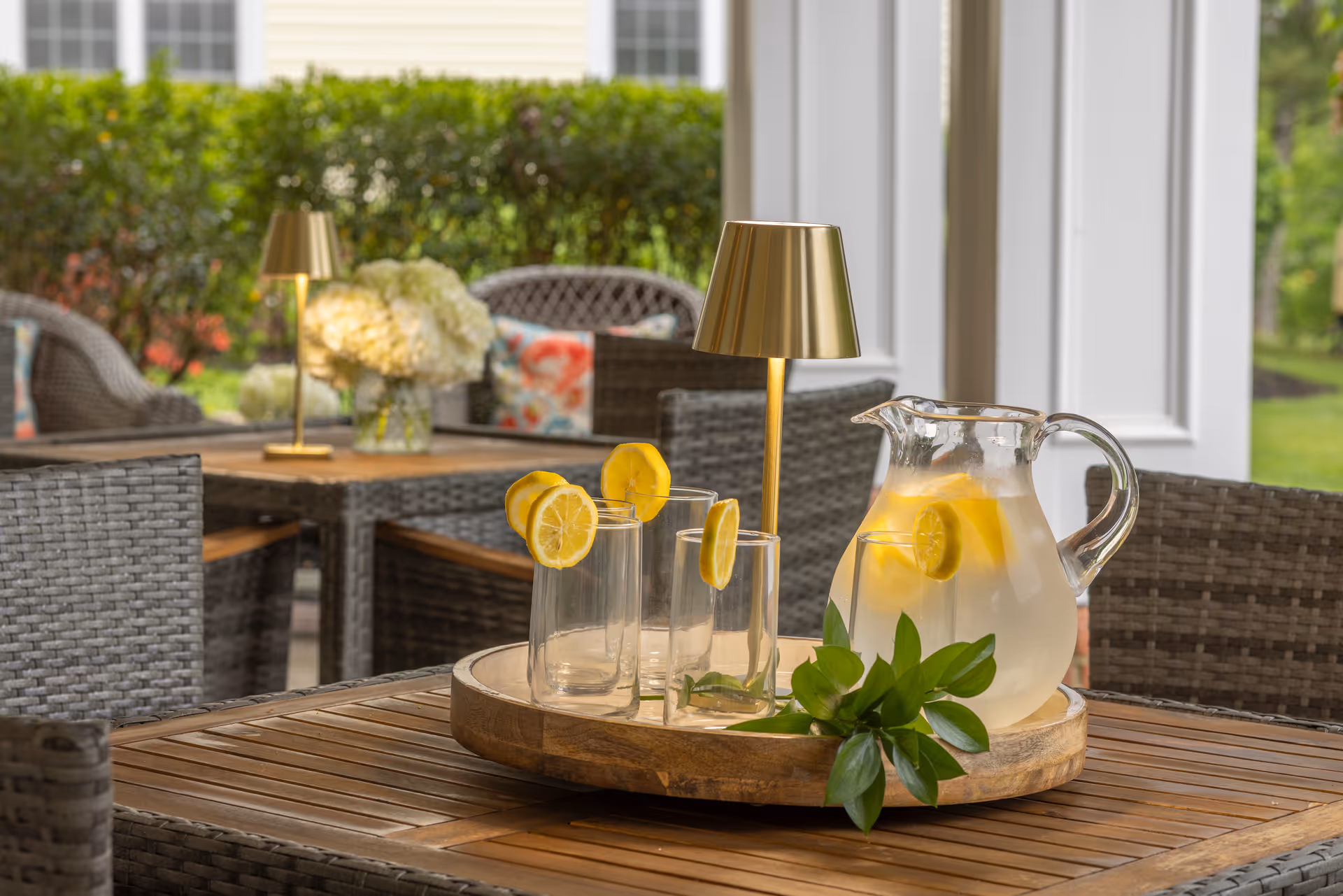 Outdoor patio area with wicker furniture and a wooden table. On the table, there is a wooden tray holding a glass pitcher of lemonade with lemon slices and three empty glasses each garnished with a lemon slice. There are two small gold-colored lamps and a vase of white flowers on a table in the background, with greenery and a white building exterior visible beyond.