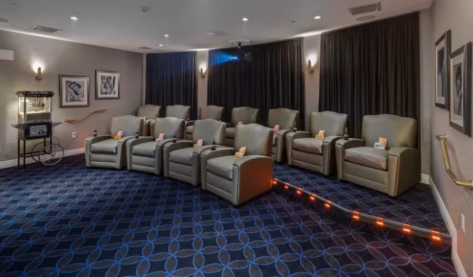 A small movie theater room with two rows of comfortable recliner chairs, each with a bottle of soda and a bag of popcorn. The room has dark curtains, patterned carpet, wall sconces, and black-and-white framed photos on the walls. A popcorn machine is visible on the left side.