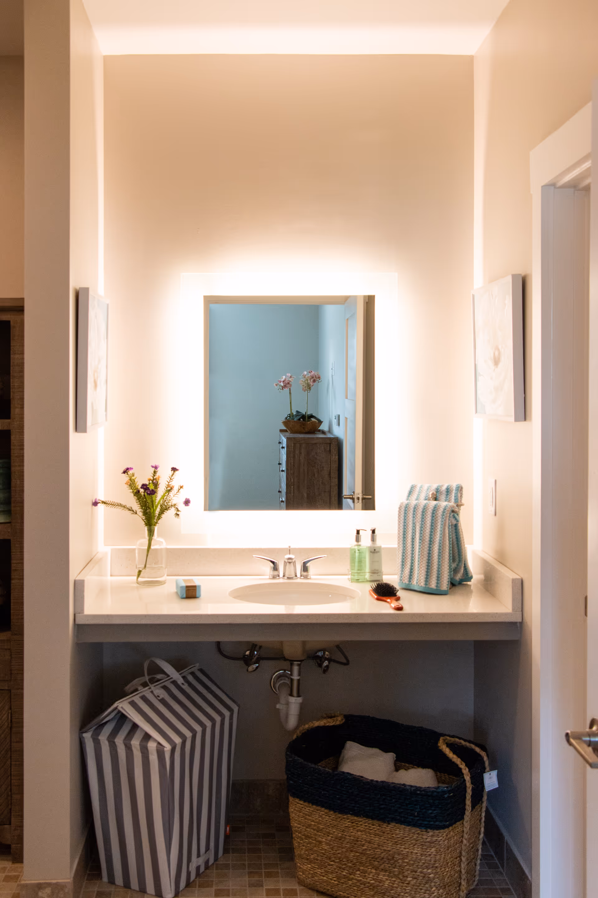 A bathroom vanity with a white countertop, an oval sink, and a modern faucet. Above the sink is a square mirror with backlighting. On the countertop are a small vase with flowers, a bar of soap, two bottles of liquid soap or lotion, a hairbrush, and folded striped towels. Below the countertop are two baskets, one striped and one woven, placed on a tiled floor. The bathroom walls are light-colored, and there are framed pictures on either side of the mirror.