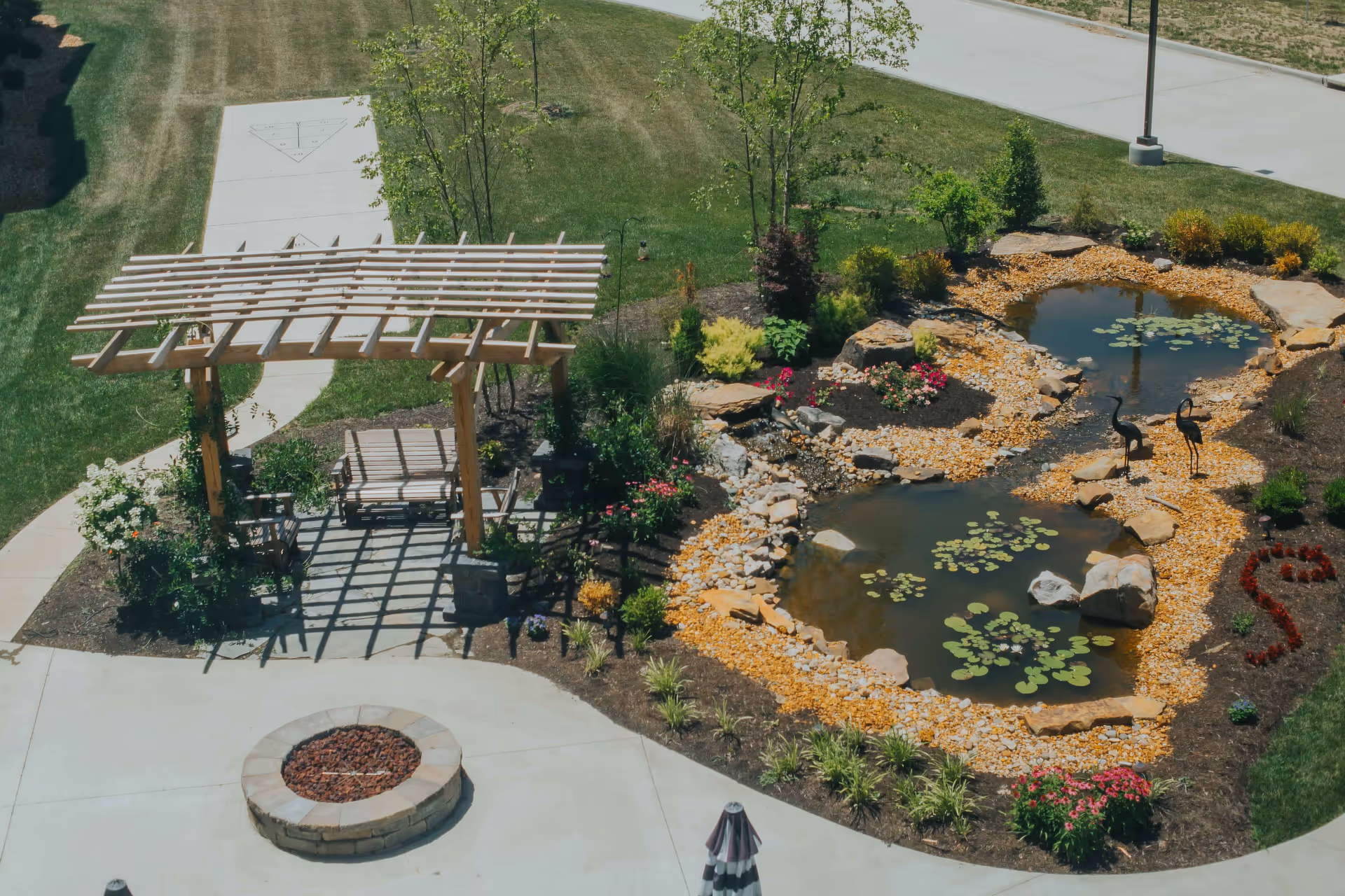 A landscaped outdoor area featuring a wooden pergola with a bench swing underneath, a small pond with lily pads and decorative rocks, two metal bird sculptures near the pond, surrounding plants and flowers, a circular stone fire pit, and paved walkways.