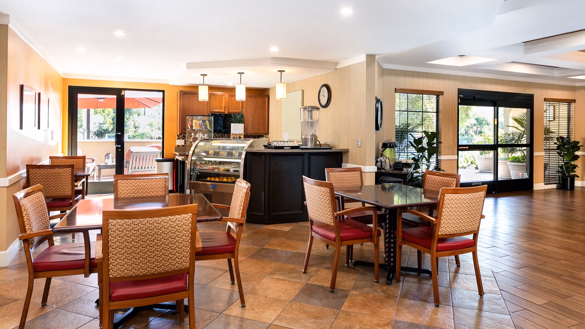 A bright and clean dining area in a senior living facility with several wooden tables and chairs with red cushions. There is a small counter with a glass display case containing food items, a water dispenser, and pendant lights hanging from the ceiling. Large windows and glass doors provide natural light and views of outdoor greenery and a patio with an umbrella.