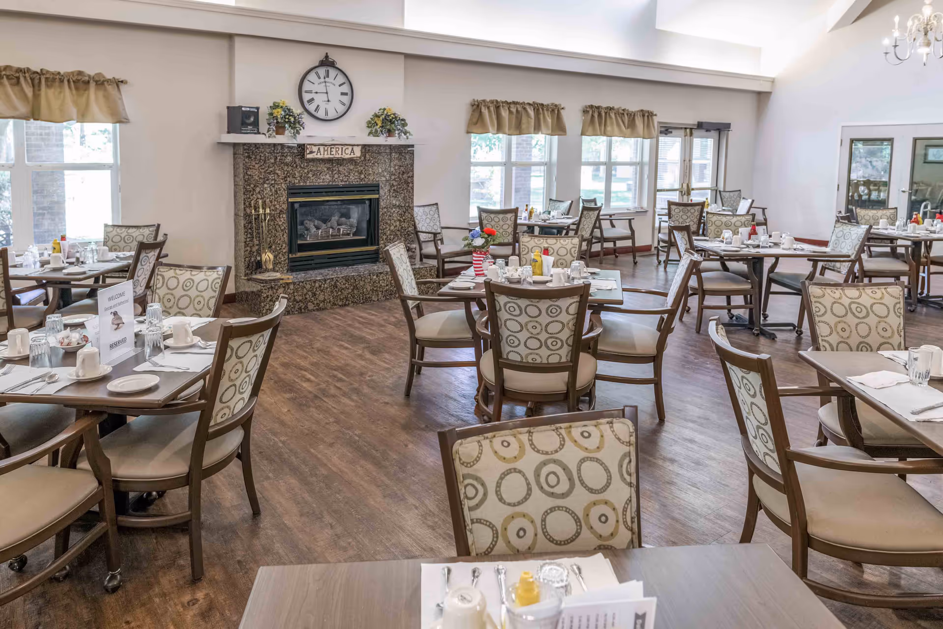 A bright dining room with multiple tables and chairs arranged neatly. Each table is set with cups, plates, utensils, and condiments. There is a fireplace with a clock above it and a sign that reads 'AMERICA'. Windows with beige curtains allow natural light to fill the room.