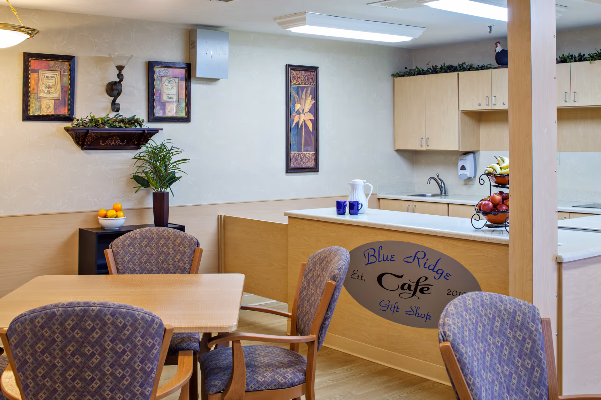 Interior view of a small cafe area named Blue Ridge Cafe within a senior living facility. The space features a wooden table with four patterned chairs, a counter with a pitcher and two blue cups, and a three-tiered fruit basket with bananas and apples. The walls are decorated with framed artwork and a small shelf with greenery. Light wood cabinetry and a sink are visible behind the counter.