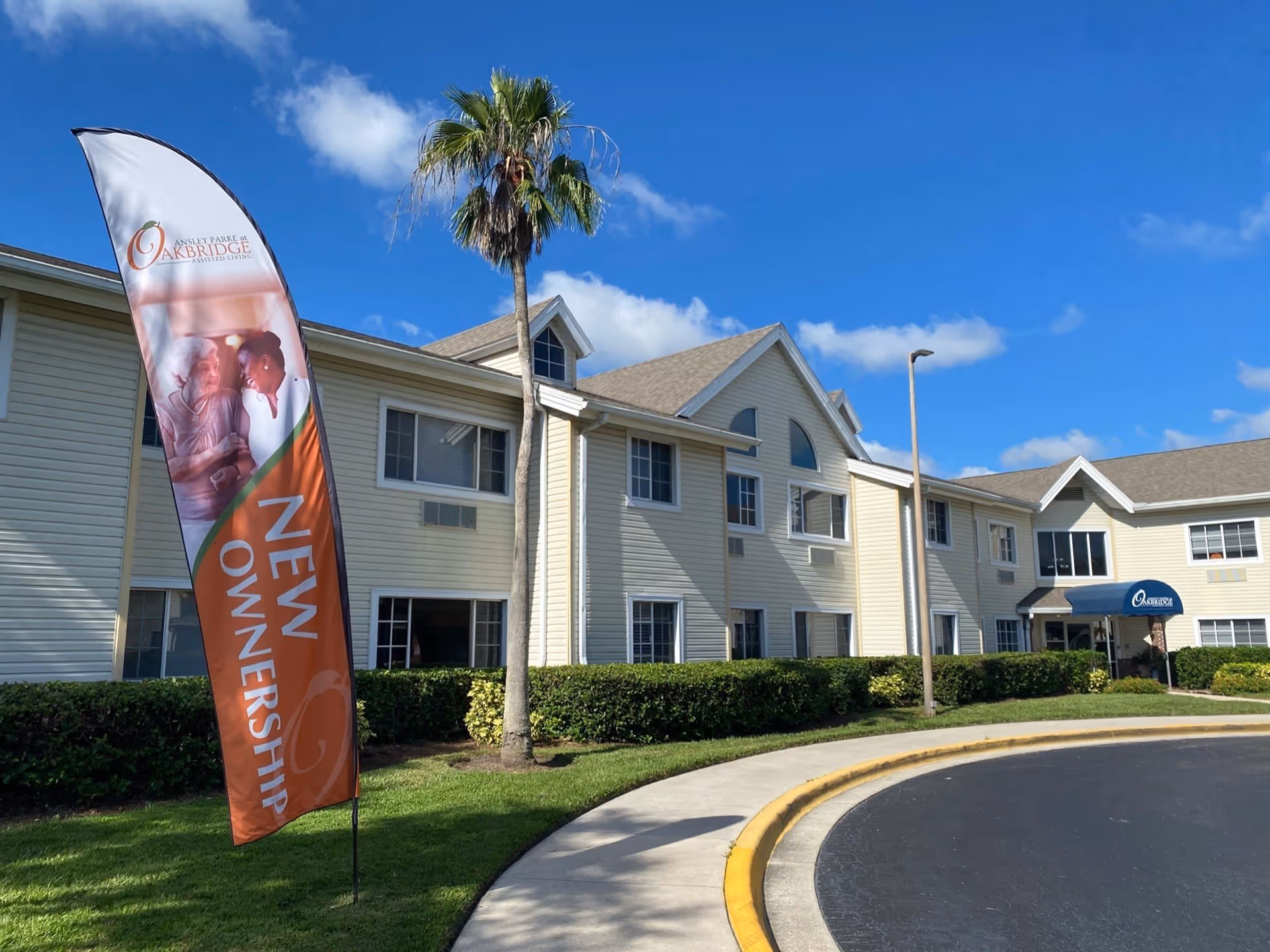 Exterior view of a two-story assisted living facility building with beige siding, several windows, and a blue awning over the entrance. A tall palm tree stands near the building, and a curved driveway with a yellow curb is in the foreground. A vertical banner flag on the lawn reads 'NEW OWNERSHIP' with the Ansley Parke at Oakbridge logo and an image of two women smiling.