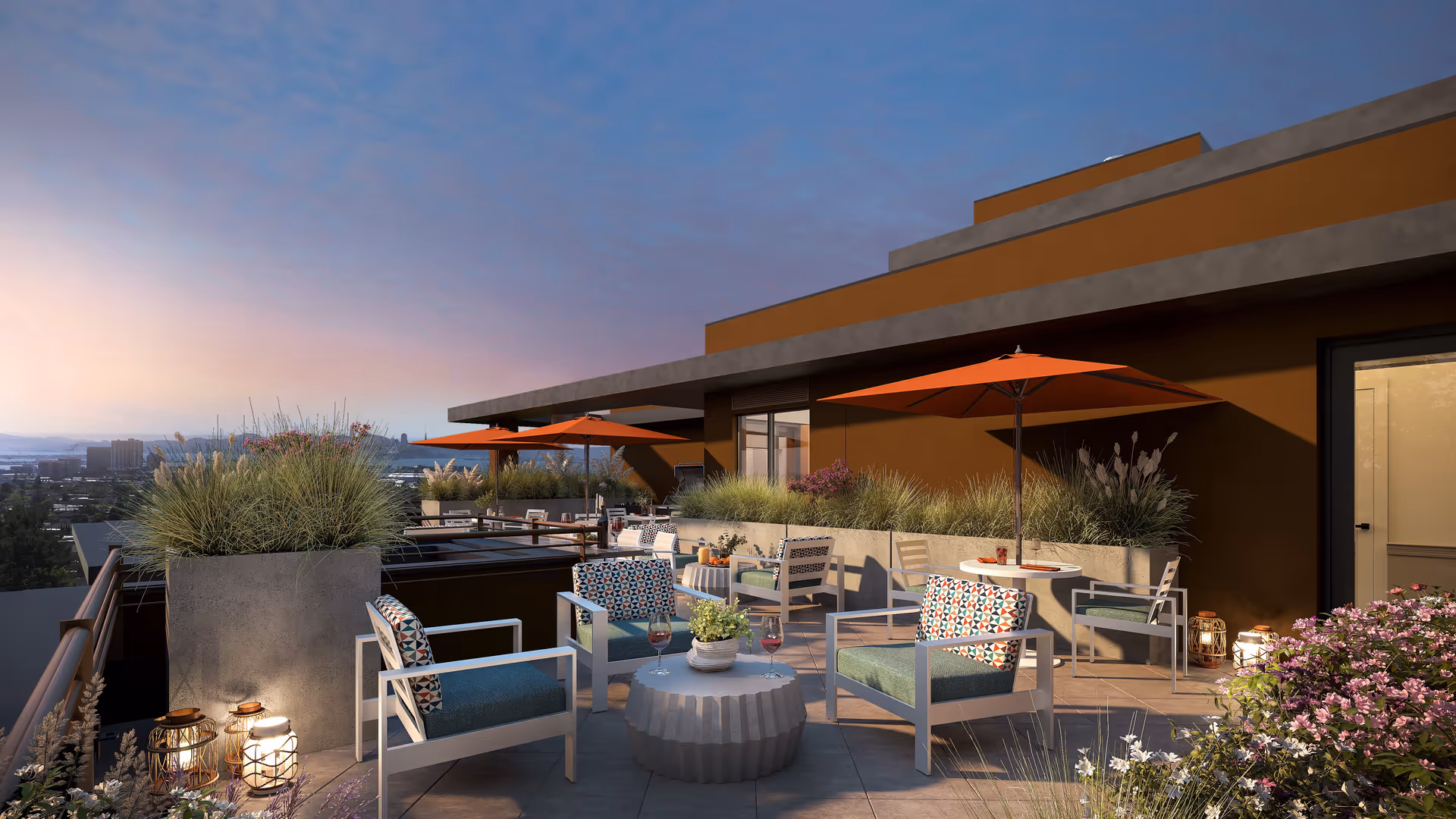 Outdoor rooftop patio area at sunset with modern seating including cushioned chairs around a small round table, orange umbrellas, potted plants, and decorative lanterns. The patio overlooks a cityscape in the distance.