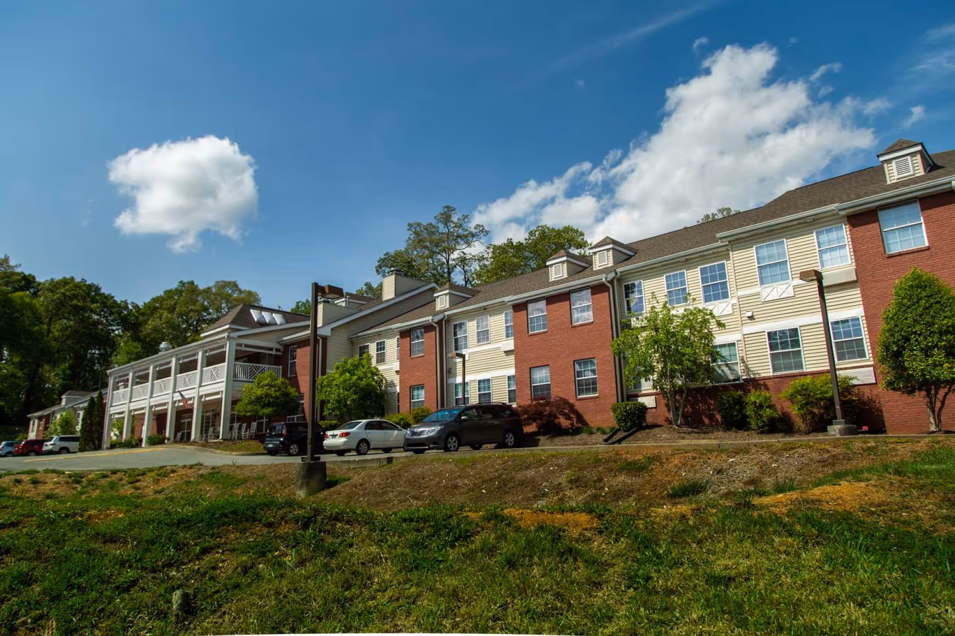 Exterior view of a two-story senior living facility building with a combination of red brick and beige siding. Several cars are parked in front of the building, which is surrounded by trees and greenery under a blue sky with some clouds.