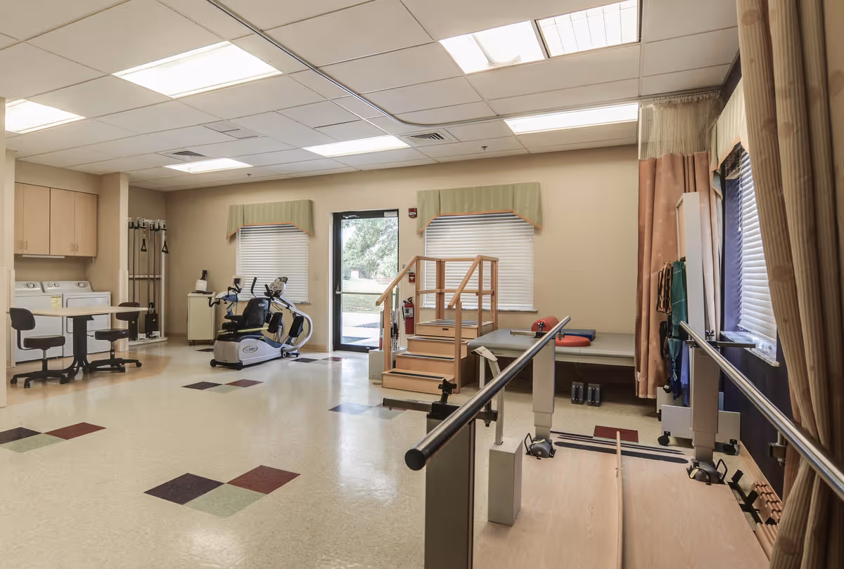 A rehabilitation or physical therapy room in a healthcare facility with exercise equipment including a recumbent bike, parallel bars for walking practice, a small set of stairs with handrails, and padded therapy tables. The room has beige walls, windows with blinds and valances, and a door leading outside. There are also cabinets and a small table with chairs.