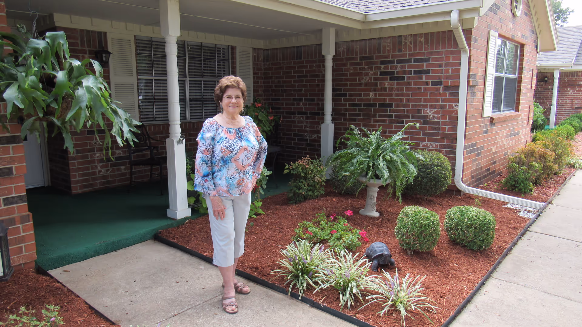 An elderly woman standing outside a brick building with a covered porch. The area features a landscaped garden bed with various plants, bushes, and a decorative turtle statue. The woman is smiling and wearing a colorful blouse and white pants.