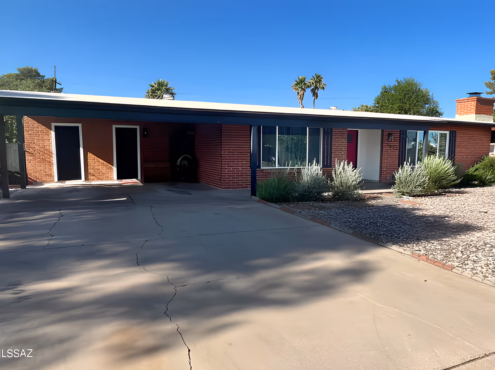 Exterior view of a single-story brick building with a flat roof and a covered carport. The building has several doors and windows, with some greenery and bushes in front. The sky is clear and blue with a few palm trees visible in the background.