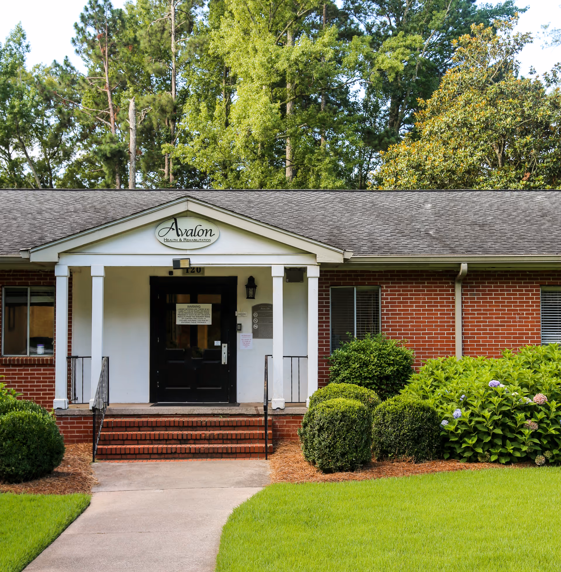 Front entrance of Avalon Health & Rehabilitation, a single-story brick building with a black door, white columns, and a small porch. The building is surrounded by neatly trimmed bushes and green grass, with tall trees in the background.