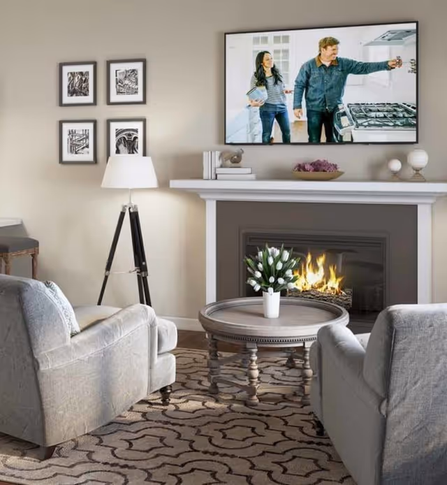 A cozy living room with two gray armchairs facing a round wooden coffee table with a white vase of flowers. Behind the table is a fireplace with a fire burning. Above the fireplace is a mounted flat-screen TV showing a man and woman in a kitchen. On the left wall are four framed black and white pictures and a tripod floor lamp.