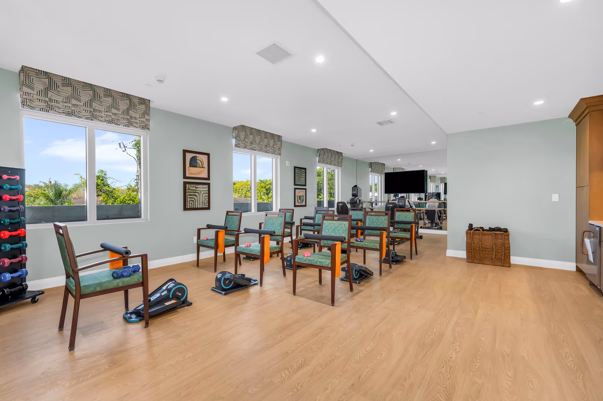 Bright fitness/activity room with rows of chairs, pedal exercisers and dumbbells in front of large windows.