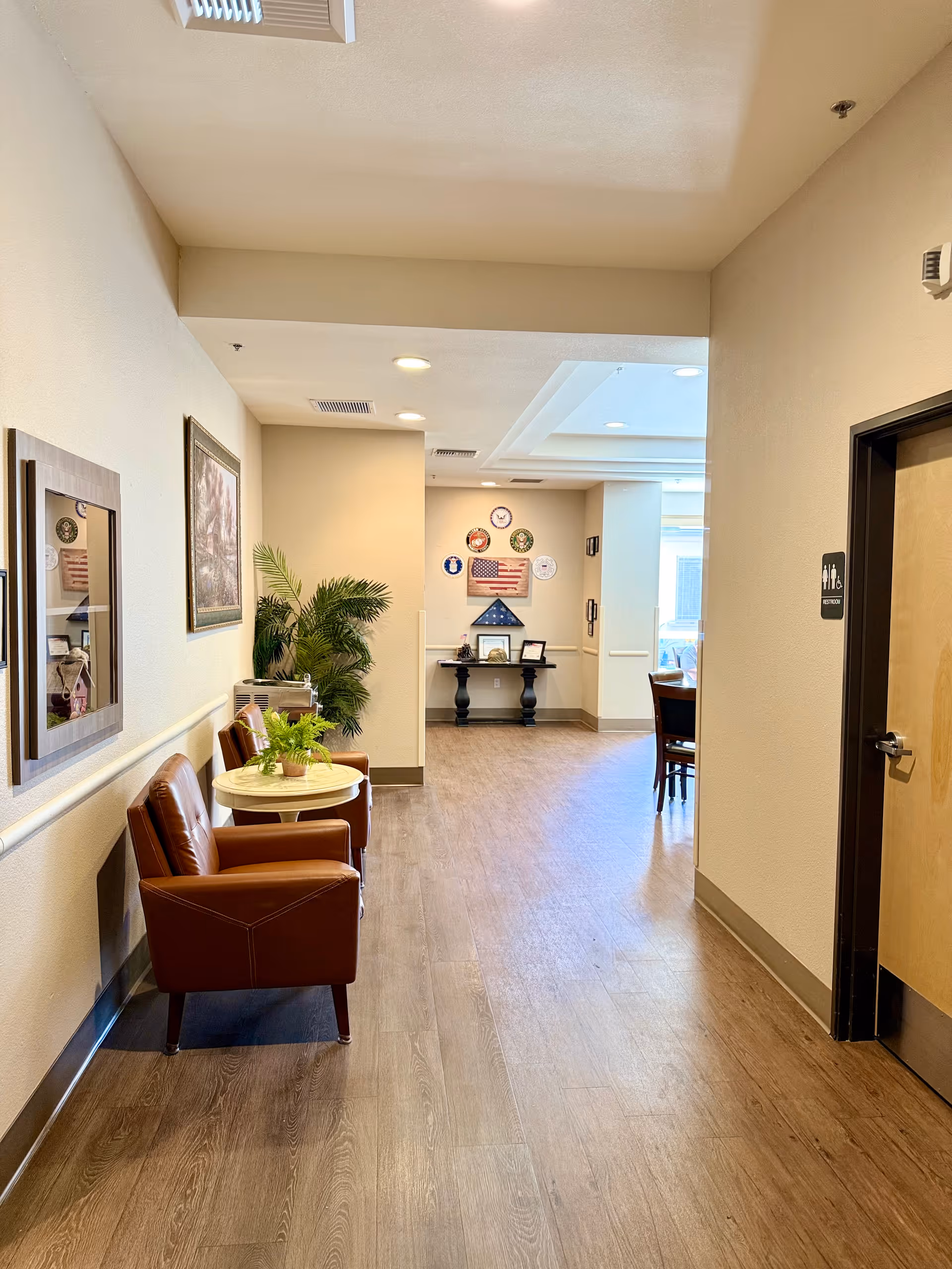 Bright interior hallway of a memory care facility with two leather chairs and a small table along a wood floor leading to a decorated wall display and dining area.