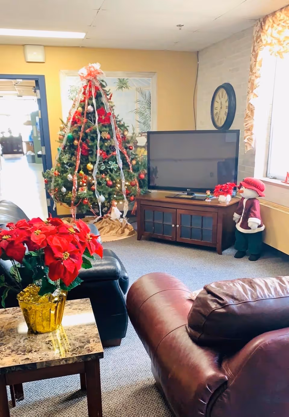 A cozy living room decorated for Christmas with a decorated Christmas tree, a TV on a wooden stand, a large clock on the wall, a poinsettia plant on a marble-top side table, and a snowman figure wearing a red hat and coat near the window.